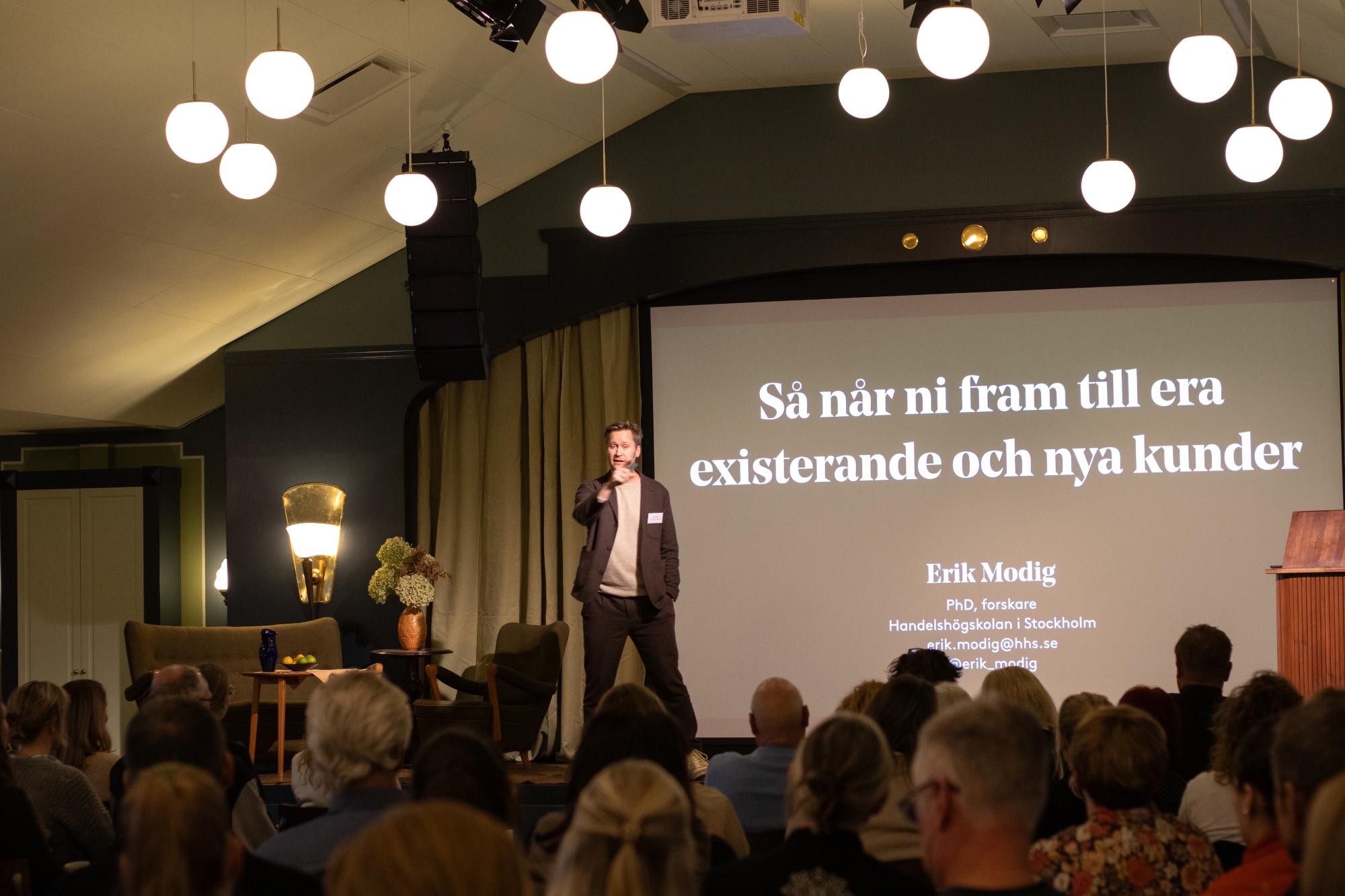 Erik Modig som står på scenen och föreläser om hur du når ut till nya och befinliga kunder. Publik runtomkring, slides bakom honom, bollformade lampor som hänger över publiken.