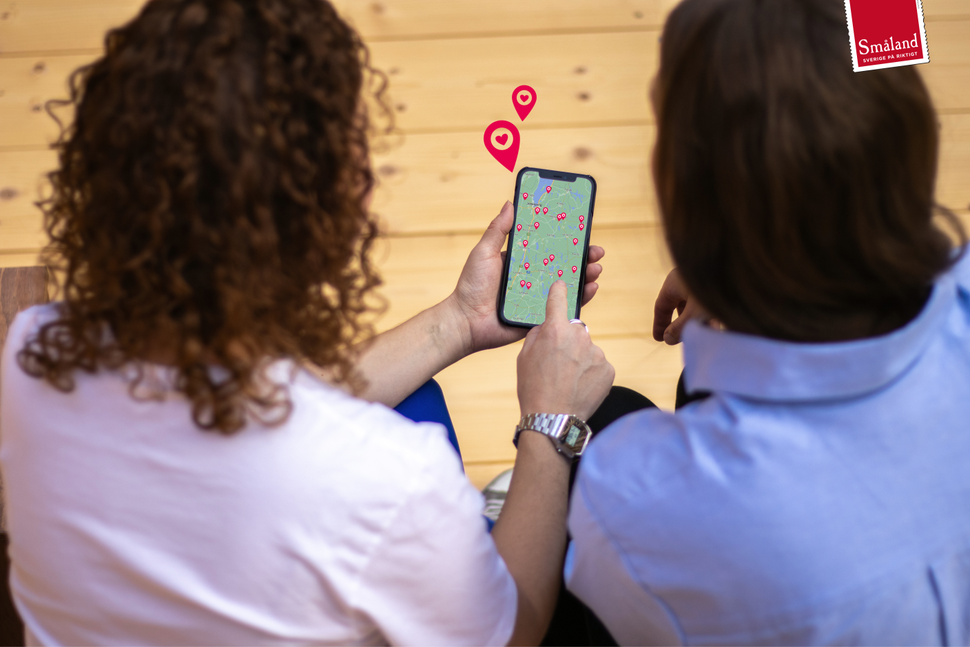 Två kvinnor med ryggen vänd mot kameran, håller upp en mobiltelefon som visar en karta med markeringar på olika destinationer.